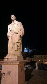 A statue at night, with a monastery illuminated on the hill in the background.